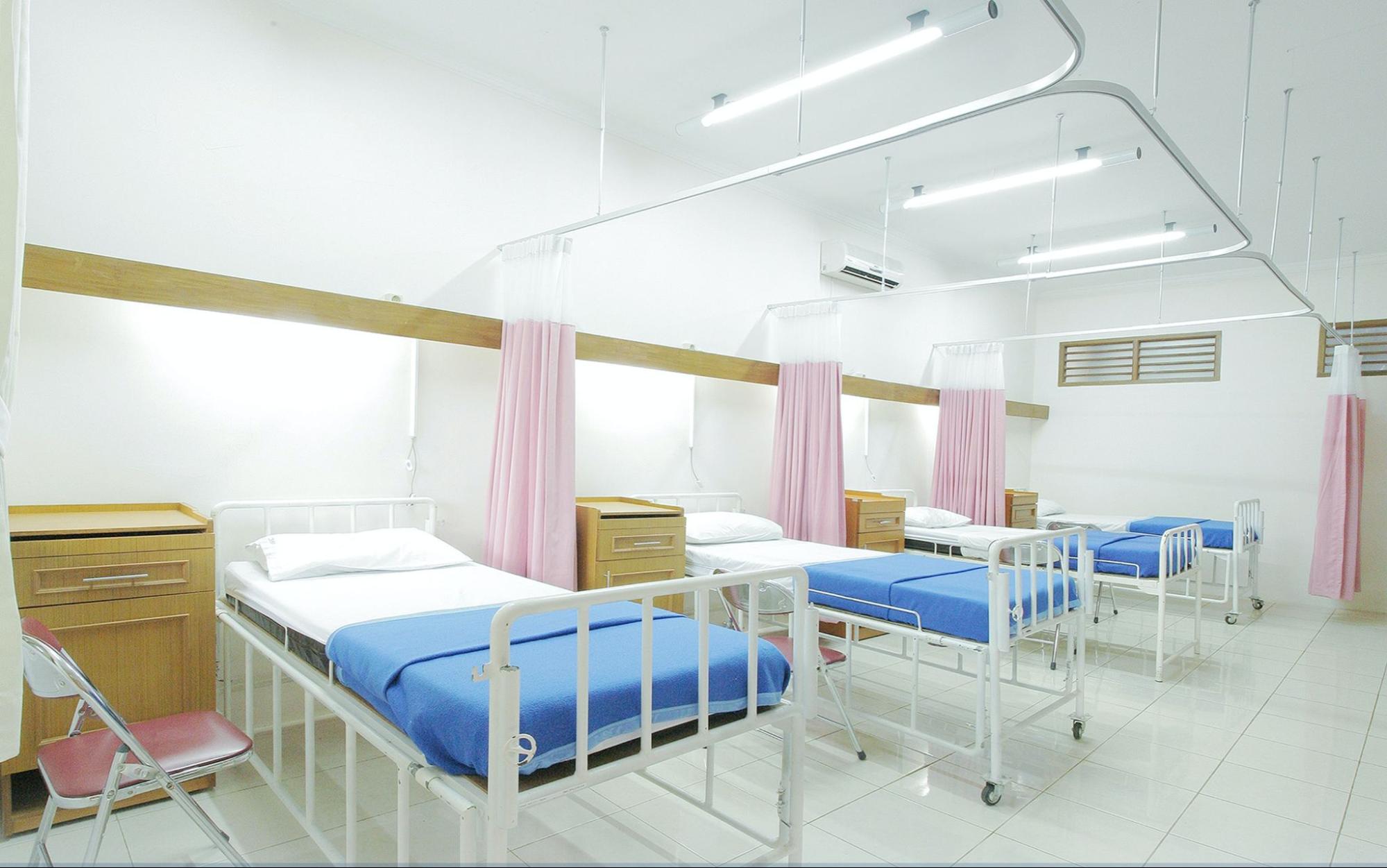 A large hospital room is filled with four hospital gurneys, with blue and white bedding, a red foldable chair to the left of each bed, a wooden side table and red privacy curtains in between each bed.