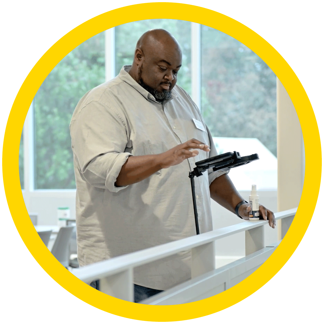 A male is standing at a railing in an office holding a solution spray bottle in his left hand, while pressing on the optisolve pathfinder ipad screen to take a photo