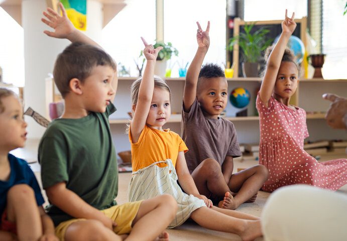 daycare facility with little children sitting beside each other raising their hands.