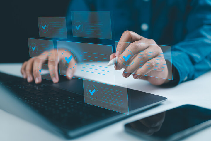A person interacts with a laptop, touching floating digital icons with check marks, symbolizing task completion or compliance verification. The scene conveys a futuristic interface with transparent overlays, enhancing the theme of digital compliance or data management. A smartphone lies nearby on the desk, adding to the tech-focused setup.