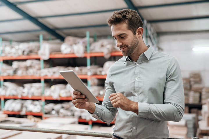 A man stands in a warehouse filled with organized shelves of materials, smiling as he checks information on a tablet. The background suggests a well-maintained inventory system, and his focused, positive demeanor implies efficient management or quality control.